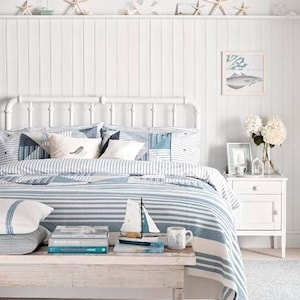 K&ouml;nnte beinhalten: Ein wei&szlig;es Bettgestell mit einer blau-wei&szlig; gestreiften Bettdecke und Kissen. Das Kopfteil ist aus Holz und wei&szlig; lackiert. An der Wand hinter dem Bett befinden sich Muscheln und Seesterne. Ein wei&szlig;er Nachttisch mit einer Schublade steht rechts neben dem Bett.