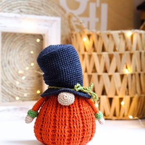 May include: A crocheted gnome with a black hat, green and yellow trim, and an orange body. The gnome is sitting on a white surface.