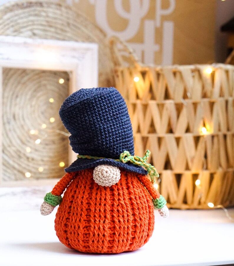 May include: A crocheted gnome with a black hat, green and yellow trim, and an orange body. The gnome is sitting on a white surface.