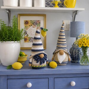May include: Two handmade crochet gnomes with striped hats in blue, beige, and white, each holding a lemon. The gnomes are on a blue surface, with a framed floral print and a potted plant in the background. Yellow lemons are placed around the gnomes.