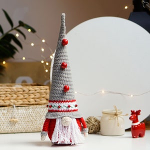 May include: A gray crocheted gnome with a red hat and white beard. The gnome has red buttons on its hat and a white and red striped band around the hat.