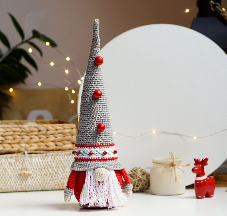 May include: A gray crocheted gnome with a red hat and white beard. The gnome has red buttons on its hat and a white and red striped band around the hat.