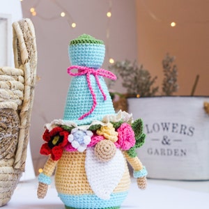 May include: A handmade crochet gnome doll with a light blue hat and body, adorned with a pink bow and a floral collar. The gnome has a white beard, a tan nose, and is decorated with red, pink, and yellow flowers. A basket and a "FLOWERS & GARDEN" sign are in the background.