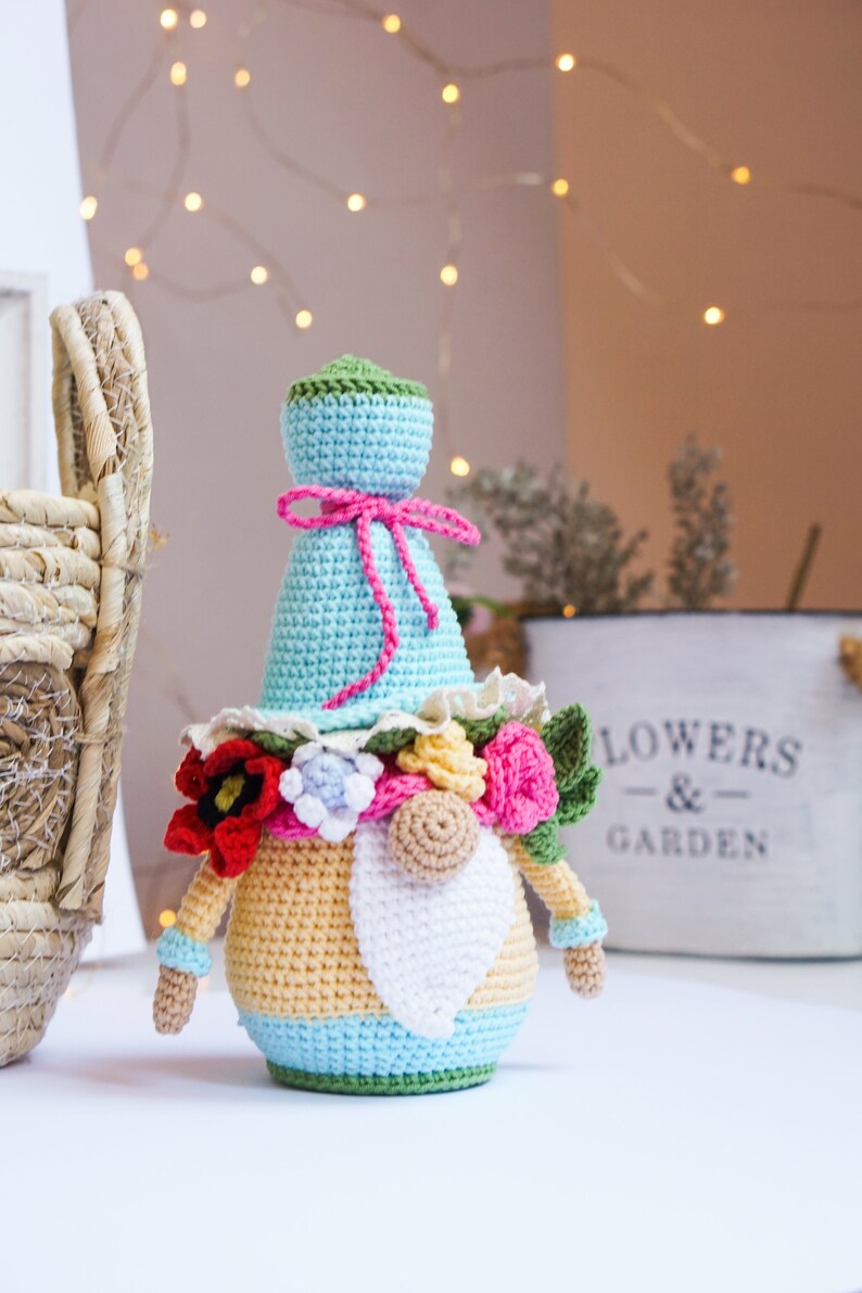 May include: A handmade crochet gnome doll with a light blue hat and body, adorned with a pink bow and a floral collar. The gnome has a white beard, a tan nose, and is decorated with red, pink, and yellow flowers. A basket and a "FLOWERS & GARDEN" sign are in the background.