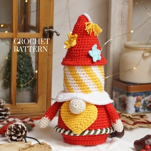 Puede incluir: Patrón de crochet para un gnomo rojo y amarillo con barba blanca y sombrero puntiagudo. El gnomo tiene un corazón amarillo en el pecho y está decorado con estrellas.