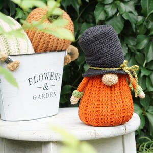 May include: A crocheted gnome with an orange body, black hat, and green accents. The gnome is sitting on a white stool in front of a metal bucket with the words "Flowers & Garden" printed on it. The bucket is filled with crocheted pumpkins.