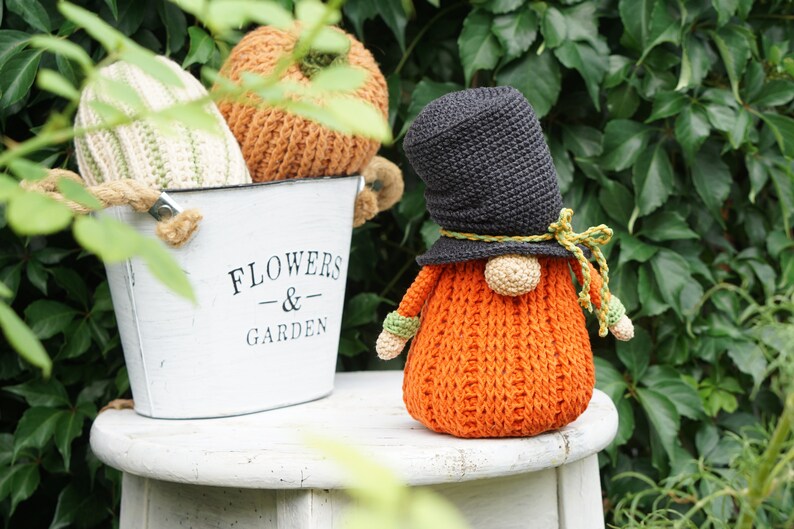 May include: A crocheted gnome with an orange body, black hat, and green accents. The gnome is sitting on a white stool in front of a metal bucket with the words "Flowers & Garden" printed on it. The bucket is filled with crocheted pumpkins.