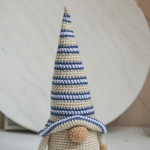 K&ouml;nnte beinhalten: Ein geh&auml;kelter Zwerg mit einer gestreiften M&uuml;tze in Blau- und Beiget&ouml;nen. Der Zwerg hat eine runde, beige Nase und einen beigefarbenen K&ouml;rper.