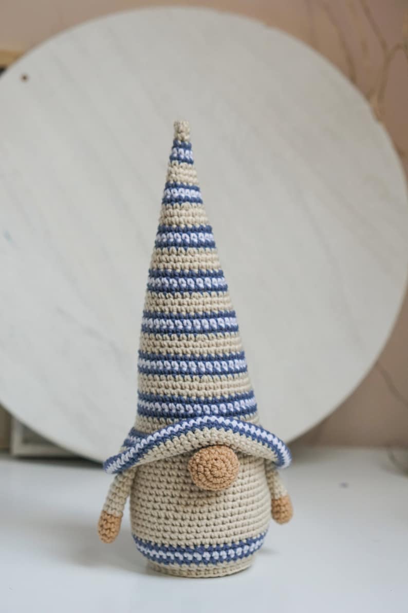 K&ouml;nnte beinhalten: Ein geh&auml;kelter Zwerg mit einer gestreiften M&uuml;tze in Blau- und Beiget&ouml;nen. Der Zwerg hat eine runde, beige Nase und einen beigefarbenen K&ouml;rper.