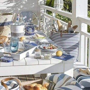 K&ouml;nnte beinhalten: Ein wei&szlig;er Tisch mit blau-wei&szlig; gestreiften Servietten, einem blau-wei&szlig; gestreiften Korb und einer Holzkiste mit dem Text "Magasin General Comptoir de Ceylan Store". Der Tisch ist mit blau-wei&szlig; gestreiften Gl&auml;sern, einem wei&szlig;en Teller mit blauem Rand und einem wei&szlig;en Teller mit blauem Rand und einer blau-wei&szlig; gestreiften Serviette gedeckt. Auf dem Tisch steht eine wei&szlig;e Kerze in einem Glasgef&auml;&szlig;.