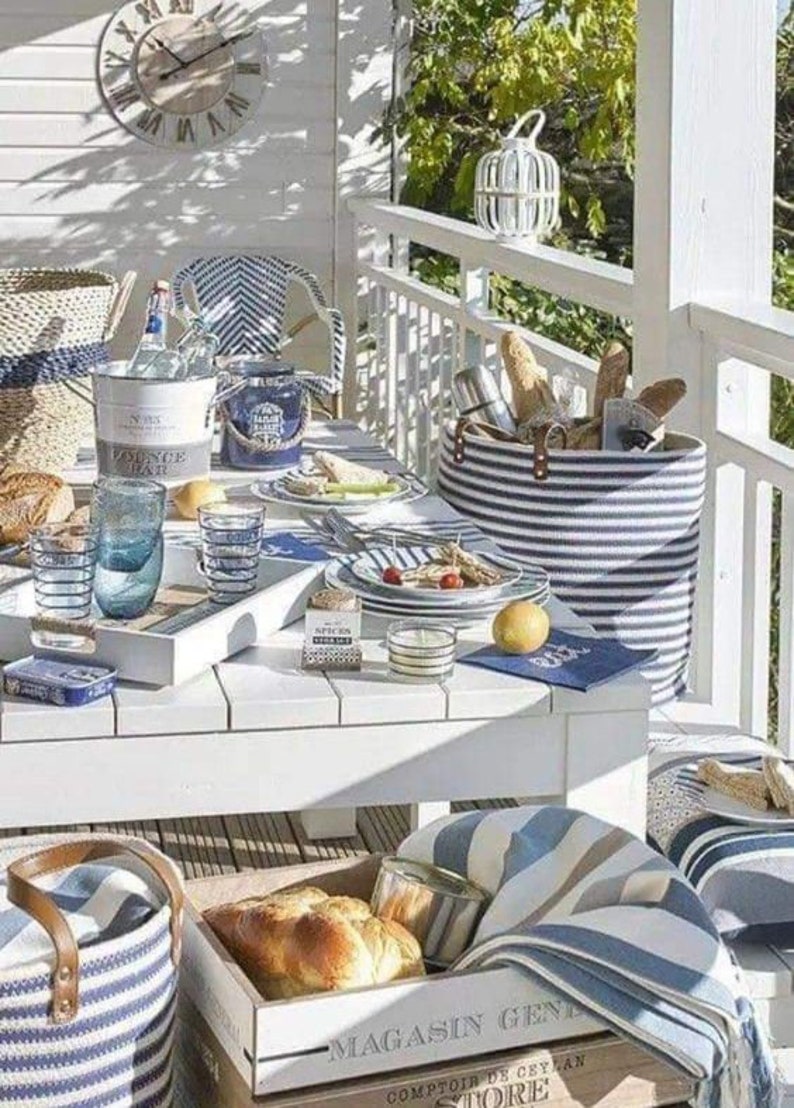 K&ouml;nnte beinhalten: Ein wei&szlig;er Tisch mit blau-wei&szlig; gestreiften Servietten, einem blau-wei&szlig; gestreiften Korb und einer Holzkiste mit dem Text "Magasin General Comptoir de Ceylan Store". Der Tisch ist mit blau-wei&szlig; gestreiften Gl&auml;sern, einem wei&szlig;en Teller mit blauem Rand und einem wei&szlig;en Teller mit blauem Rand und einer blau-wei&szlig; gestreiften Serviette gedeckt. Auf dem Tisch steht eine wei&szlig;e Kerze in einem Glasgef&auml;&szlig;.