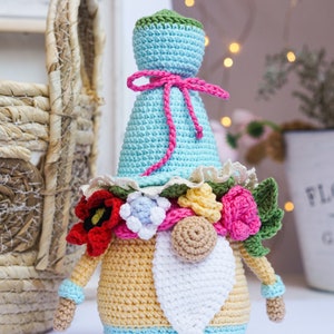May include: Crocheted gnome doll with a light blue hat and body, adorned with a pink bow and a floral garland. The gnome has a white beard, tan arms, and is approximately 6 inches tall. A whimsical, handmade decorative item.