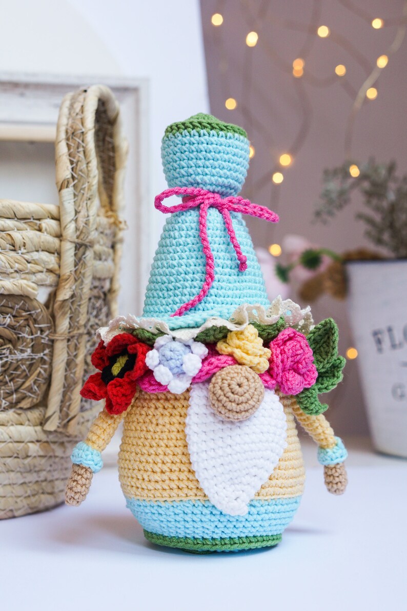 May include: Crocheted gnome doll with a light blue hat and body, adorned with a pink bow and a floral garland. The gnome has a white beard, tan arms, and is approximately 6 inches tall. A whimsical, handmade decorative item.