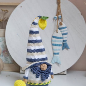 May include: Crocheted gnome figurine with a long, striped hat and a yellow lemon. The gnome has a white body, blue beard, and a small, round nose. A second lemon is next to the gnome. Decorative fish hang in the background.