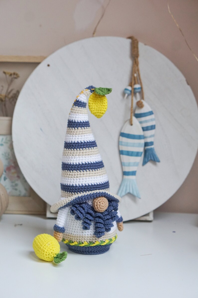May include: Crocheted gnome figurine with a long, striped hat and a yellow lemon. The gnome has a white body, blue beard, and a small, round nose. A second lemon is next to the gnome. Decorative fish hang in the background.