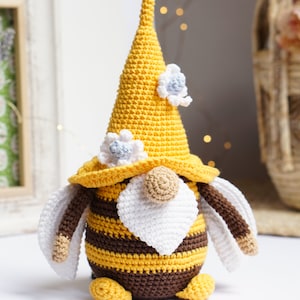 Könnte beinhalten: Ein gehäkelter Zwerg, der ein gelbes und braunes Bienenkostüm mit weißen Flügeln und einem weißen Bart trägt. Der Zwerg trägt einen gelben Hut mit weißen Blumen.