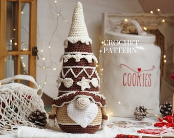 May include: Crocheted gingerbread gnome ornament with a white beard and a heart-shaped nose. The gnome is wearing a brown hat with white icing and a brown and white patterned sweater. The gnome is sitting on a white surface with red berries and a white crocheted bag in the background.