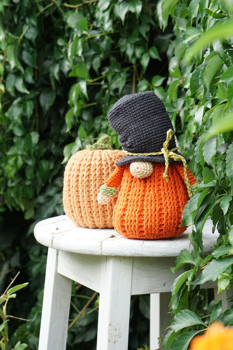 May include: A crocheted gnome wearing a black hat and holding a small orange pumpkin. The gnome and pumpkin are sitting on a white stool in front of a green leafy background.