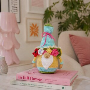 May include: A handmade crochet gnome doll with a blue hat tied with a pink bow. The gnome has a yellow body, a white beard, and is adorned with colorful flowers. It sits atop a stack of pink books with the title "In full flower".