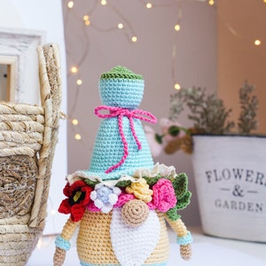 May include: A handmade crochet gnome doll with a light blue hat and body, adorned with a pink bow and colorful floral accents. The gnome has a white beard and is approximately 7 inches tall, perfect for home decor.