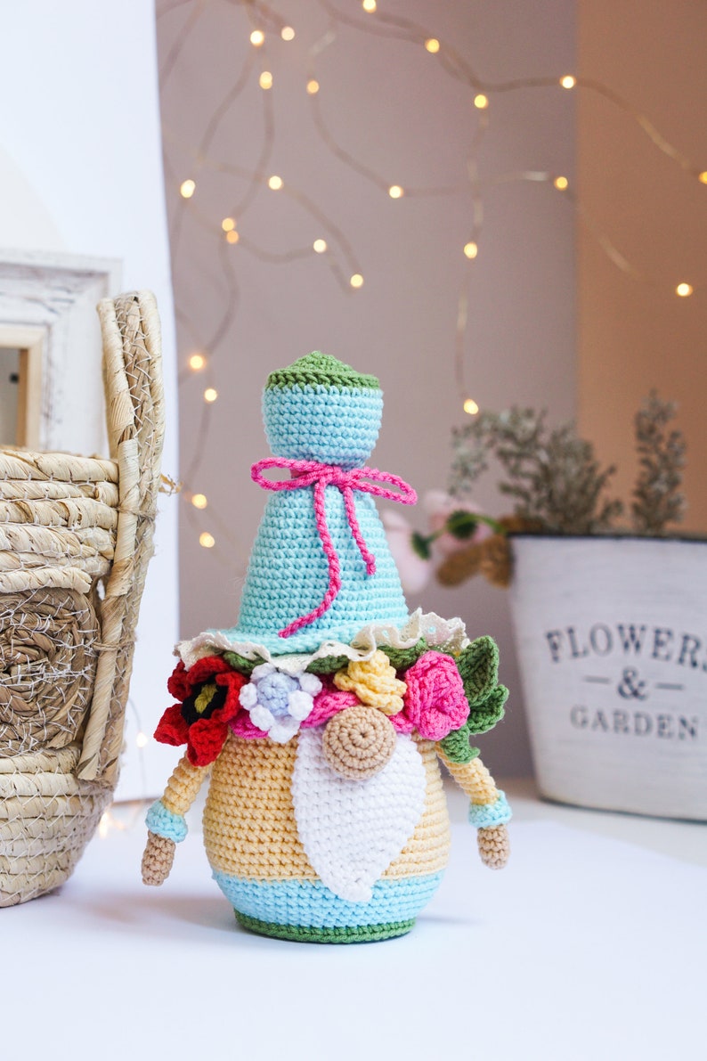 May include: A handmade crochet gnome doll with a light blue hat and body, adorned with a pink bow and colorful floral accents. The gnome has a white beard and is approximately 7 inches tall, perfect for home decor.