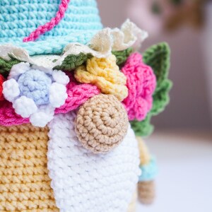May include: Crocheted gnome doll with a light blue hat and a white beard. The gnome is adorned with colorful crocheted flowers in pink, yellow, and blue. A pink cord is attached to the hat. The gnome's body is yellow and blue.