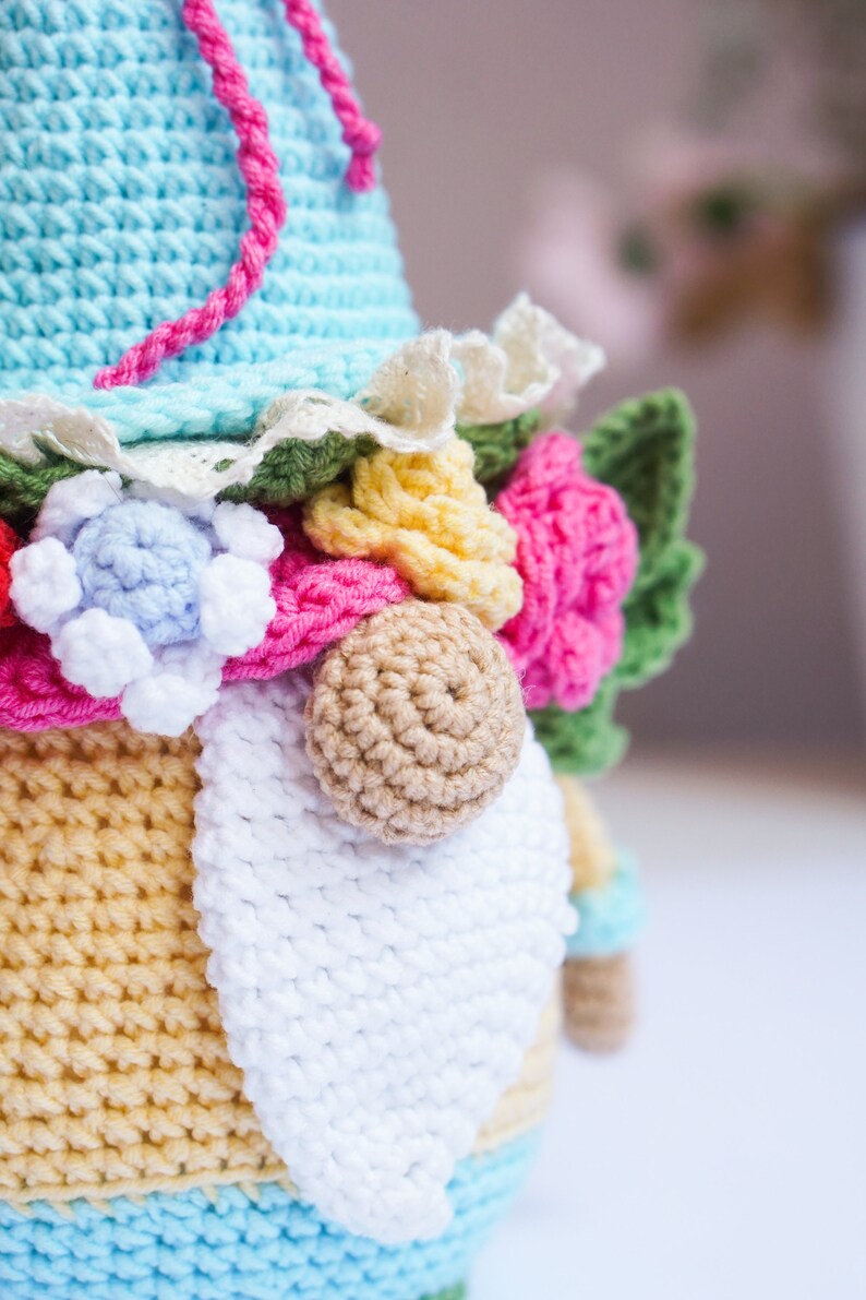 May include: Crocheted gnome doll with a light blue hat and a white beard. The gnome is adorned with colorful crocheted flowers in pink, yellow, and blue. A pink cord is attached to the hat. The gnome's body is yellow and blue.