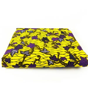 Op de afbeelding: Gevouwen stof met een heldergele achtergrond en een herhalend patroon van paarse en witte bladeren verbonden door bruine wijnranken. De stof heeft een rasterachtige textuur.