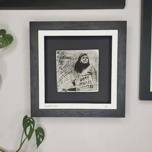 Op de afbeelding: Een ingelijste zwart-witfoto van een vrouw die een bord vasthoudt met de tekst "Vote Peoples Democrac For All". De foto is gemonteerd op een witte passe-partout en ingelijst in een zwart frame.