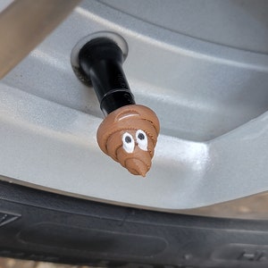 Könnte beinhalten: Eine braune, neuartige Reifenventil-Kappe in Form eines lächelnden Kackhaufen-Emojis. Die Kappe ist an einem schwarzen Ventil an einem silbernen Autorad befestigt, mit einem schwarzen Reifen sichtbar.