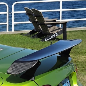 May include: A green sports car with a black spoiler is parked on a grassy area near a body of water. The spoiler has a carbon fiber texture. Two wooden chairs are in the background, and a white railing is visible.