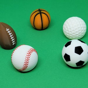 May include: An assortment of miniature sports balls, including a brown football, an orange basketball, a white golf ball, a white baseball with red stitching, and a black and white soccer ball, all set against a green background.