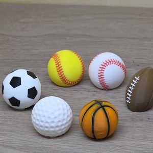 Puede incluir: Una colección de cinco pelotas deportivas en miniatura que incluye un balón de fútbol blanco y negro, una pelota de sóftbol amarilla, una pelota de béisbol blanca con costuras rojas, una pelota de golf blanca con hoyuelos, un balón de baloncesto naranja y un balón de fútbol americano marrón con cordones blancos.