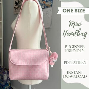 Puede incluir: Un mini bolso de mano con estampado de cuadros vichy en rosa claro y blanco, con cierre de solapa y una correa larga para el hombro. Un pequeño dije de peluche rosa con un detalle de flor blanca cuelga del lateral mediante un clip.