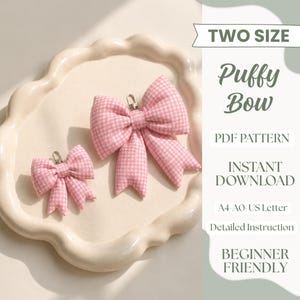 Könnte beinhalten: Zwei rosa-weiß karierte Stoffschleifen in verschiedenen Größen, jeweils mit einem kleinen Metallkarabiner an der Oberseite. Der Text lautet: TWO SIZE Puffy Bow PDF PATTERN INSTANT DOWNLOAD A4-A0-US Letter Detailed Instruction BEGINNER FRIENDLY.