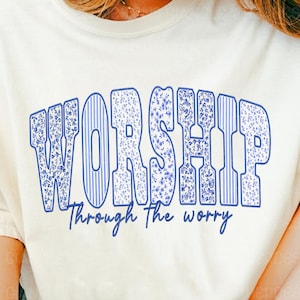 Puede incluir: Camiseta color crema con la palabra "WORSHIP" en letras azules con estampado floral. Debajo está la frase "Through the worry". La camiseta es de estilo casual.
