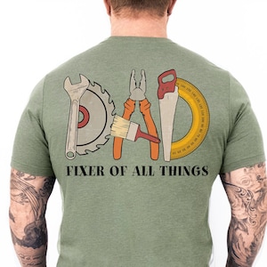 Puede incluir: Camiseta verde con un gráfico de herramientas que deletrean "DAD" y el texto "Fixer of All Things".
