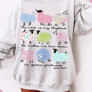 May include: A light grey sweatshirt with a design of colourful sheep illustrations and the text "The Lord is my Shepherd" and "He makes me lies down in green pastures" from Psalm 23:1-2. The sheep are in various colours and patterns.