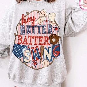 Puede incluir: Sudadera gris claro con un diseño temático de béisbol. El gráfico incluye las palabras "Hey Batter Batter Swing" en rojo y azul, junto con elementos de béisbol como un bate, una pelota y un guante. Un lazo y estrellas completan el diseño.