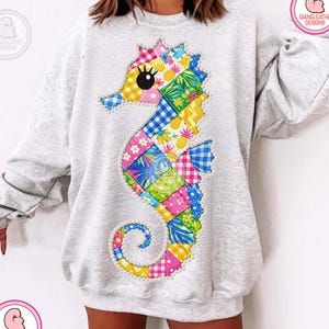 May include: A light gray sweatshirt featuring a colorful seahorse design. The seahorse is made of patchwork squares in various patterns and colors, including pink, blue, yellow, and green. The design includes the text "GIANGLEATHE DESIGNS" in a pink circle.