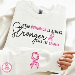 Könnte beinhalten: Weißer Pullover mit dem Text "The Comeback is Always Stronger Than the Setback" und einem rosa Band aus Herzen auf der Vorderseite. Der linke Ärmel hat ein rosa Band aus Herzen.