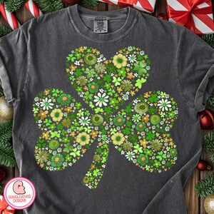 May include: A dark gray t-shirt featuring a large shamrock design filled with various green, yellow, and white flowers. The shamrock is heart-shaped. The shirt has a crew neck and short sleeves.