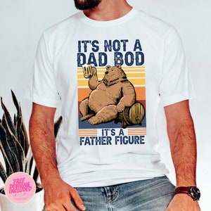May include: White t-shirt with a retro-style graphic of a bear holding a beer and the text "It's not a dad bod, it's a father figure."