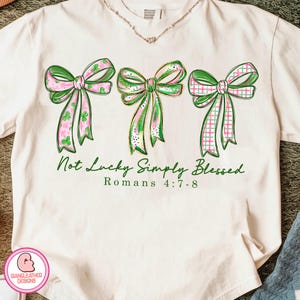 以下が含まれることがあります： クリーム色のTシャツ。緑とピンクの3つのリボンが描かれており、それぞれ異なる模様です。「Not Lucky Simply Blessed」と「Romans 4:7-8」の文字が緑色でリボンの下にプリントされています。左下にはロゴがあります。