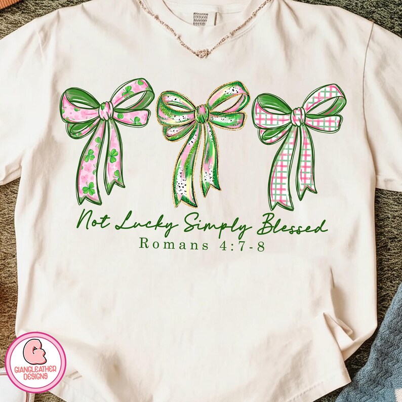以下が含まれることがあります： クリーム色のTシャツ。緑とピンクの3つのリボンが描かれており、それぞれ異なる模様です。「Not Lucky Simply Blessed」と「Romans 4:7-8」の文字が緑色でリボンの下にプリントされています。左下にはロゴがあります。