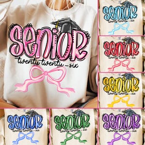 Könnte beinhalten: Ein cremefarbenes Sweatshirt mit dem Wort "Senior" in Rosa mit schwarzer Umrandung, einer Abschlusskappe und dem Text "twenty twenty-six". Unter dem Text befindet sich eine rosa Schleife. Weitere Shirts mit dem gleichen Design in verschiedenen Farben sind ebenfalls abgebildet.