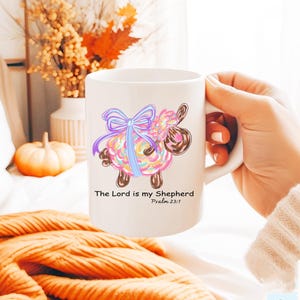 May include: White ceramic mug with a colorful cartoon sheep with a blue bow. The mug has the text "The Lord is my Shepherd" and "Psalm 23:1" printed on it.