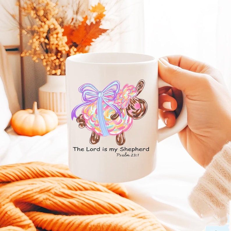 May include: White ceramic mug with a colorful cartoon sheep with a blue bow. The mug has the text "The Lord is my Shepherd" and "Psalm 23:1" printed on it.