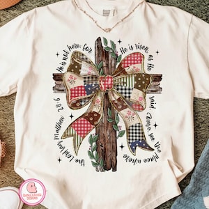 May include: Cream-colored t-shirt featuring a cross design with a patchwork bow. The cross is made of wood, and the bow has various patterns. Text around the cross reads "He is not here: for He is risen, as He said. Come, see the place where The Lord lay Matthew 28:6."