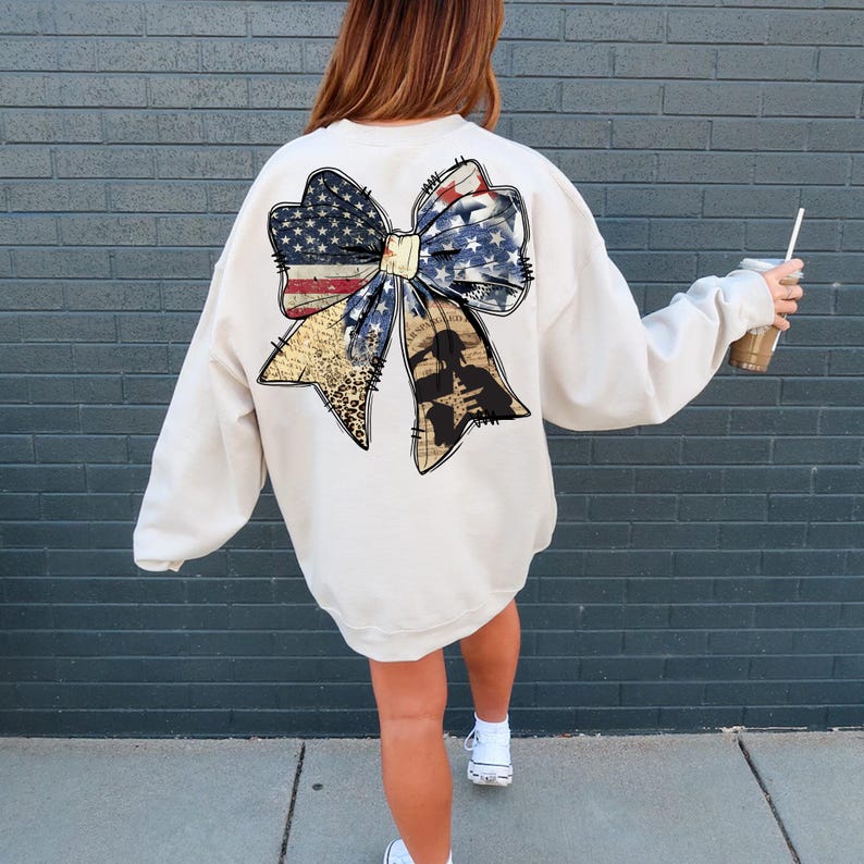 May include: A cream-colored sweatshirt with a large, distressed American flag bow on the back. The bow is made of fabric with a vintage look and features a silhouette of a soldier in the center.